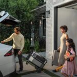 A family loads a large suitcase and a rolling cart next to a white car in a driveway, with a woman and a girl carrying luggage.