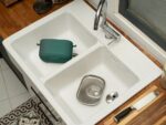 White double kitchen sink with a chrome faucet, a green silicone dish container resting in the left basin, a metal tray in the right basin, and a chef's knife on the wooden counter nearby.
