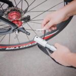 Person inflating a bicycle tire with a handheld pump, attaching the nozzle to the valve near the rear wheel hub.]