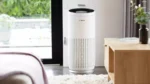Tall white Bosch air purifier with vertical slits stands in a living room near a window and wooden table behind it.
