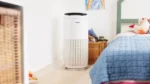 White Bosch tower air purifier with a black top vent beside a blue quilted bed in a bright kid's bedroom, with a toy basket and rainbow artwork nearby.