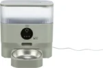 Automatic pet feeder with a translucent hopper and a circular control panel, beige base, feeding bowl in front, and a power cord to the side (Trixie brand).