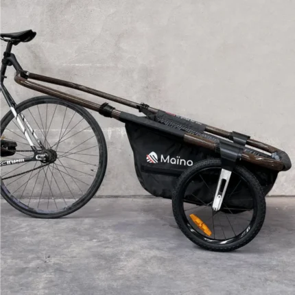 A bicycle with a long wooden rack pulling a black Maino cargo trailer bag attached to a front wheel, set against a gray wall.