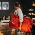 Man carries a large red Croozer bag over his shoulder in a modern kitchen.