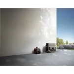 Red portable generator and a gray power storage unit connected by cords inside a bright, minimalist room; solar panels visible outside.