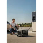 Man crouches beside a portable power station connected to an electric car at an outdoor charging station on a sunny day.