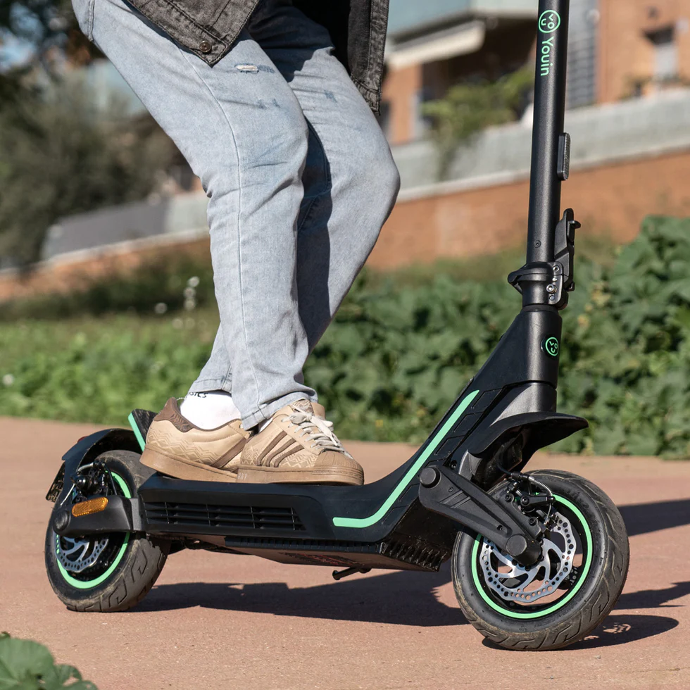 Feet and lower legs of a rider on a black electric scooter with green accents, wearing light jeans and beige sneakers on a paved path.