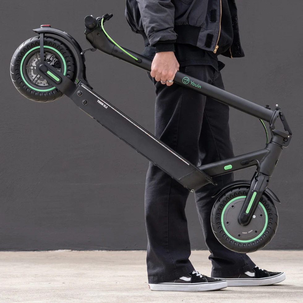 Person in dark clothing carrying a folded black electric scooter with green accents and large wheels.