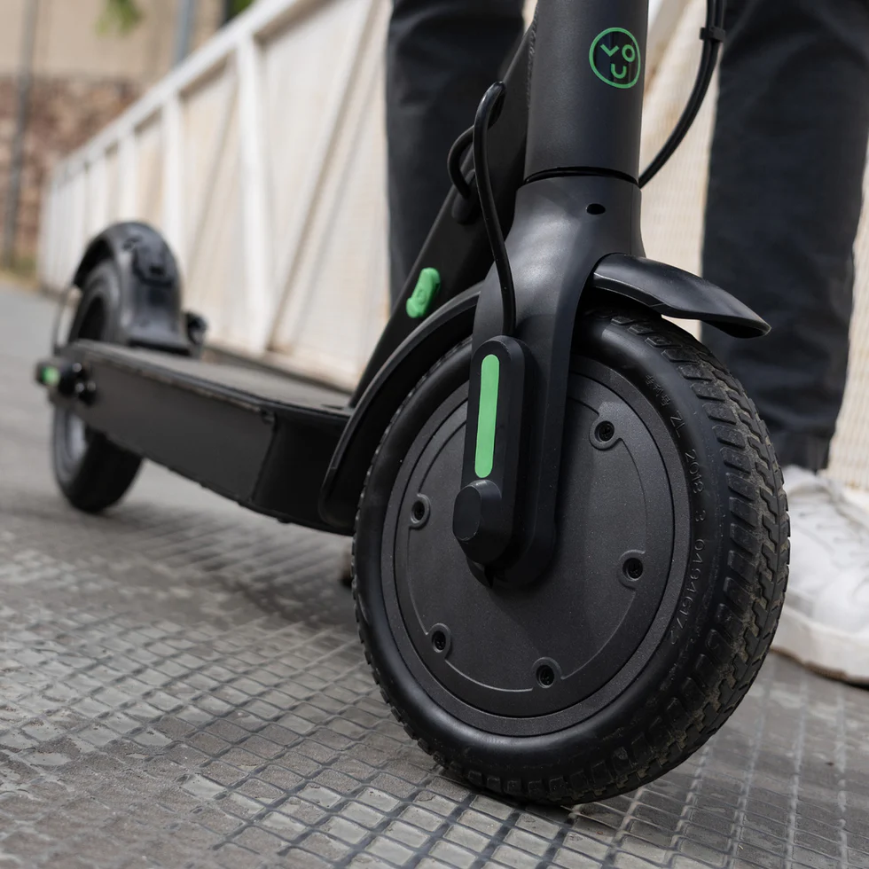 Front wheel of an electric scooter with black tire and green accent on the fork, on a sidewalk curbside.