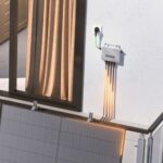 Outdoor wall with an electrical junction box and five orange cables running down to a solar panel installation beneath a window with beige curtains