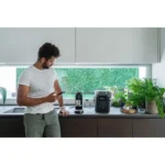 Man in a white t-shirt leans on a kitchen counter, looking at his phone beside a coffee machine and grinder with plants nearby.
