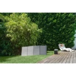 Backyard with bamboo and a dense hedge, four gray foldable panels on the grass, and a person sunbathing on a lounge chair by a small speaker on a wooden deck.