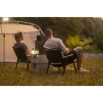 Two people sit in camping chairs beside a tent and lantern, relaxing in a grassy outdoor area at dusk.