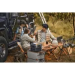 Family campsite scene: three people chat and laugh near a SUV, with a table, kettle, and portable stove set up on gear-filled ground