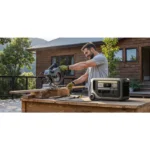 Man wearing gloves uses a miter saw on a wooden workbench outdoors, with a portable generator nearby.
