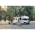 RV parked on a forest road with a large portable solar panel set up; two people sit in camping chairs nearby.
