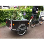 Bike with a plant-filled trailer attached on a dirt path, person riding nearby in black jacket.