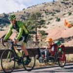 Cyclist in green gear rides a road bike while towing a two-child trailer with helmets along a hillside road; orange trailer flag visible.