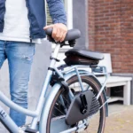 Person in a denim jacket and jeans holds the saddle of a light blue electric bicycle on a sidewalk.