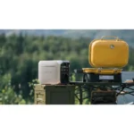 White portable generator sits on a green ammo tin next to a bright yellow hard-shell case on a camping table outdoors with trees in the background.