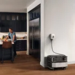 A man leaning on a kitchen island with fruit basket, while a compact wheeled home robot sits plugged into the wall nearby.