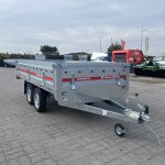 Silver open utility trailer with tandem wheels and a hitch, parked on a paved lot with other vehicles nearby.