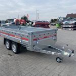 Twin-axle open utility trailer with a hitch, parked on a paved lot, with trucks in the background.