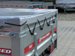 Close-up of a silver metal trailer with a grey cover, black bungee cords along the side, and a red TEMARED label.
