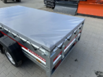 Red and gray utility trailer with a gray tarp cover secured by bungee cords on a paved lot.