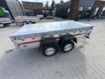 Dual-axle aluminum utility trailer with a gray cover secured by cords, featuring a red stripe and parked on a paved lot.