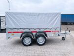 Side view of a dual-axle trailer with a white tarpaulin cover and red stripe, parked on a paved lot with a blue trailer in the background