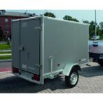 Grey enclosed cargo trailer hitched to a vehicle, viewed from the rear-left, on a paved parking lot.
