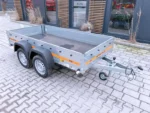 Two-axle grey metal utility trailer with a flatbed and orange safety stripe, hitched to a tow bar, parked outside a modern building.
