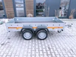 Twin-axle gray utility trailer with an orange stripe, parked on a brick surface in front of a modern building; hitch visible at the front and wheels covered by black fenders.