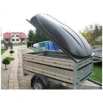Small utility trailer with a hinged lid open, parked on a driveway in front of a yard and house area.