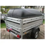 Silver Brenderup single-axle cargo trailer with a black hard-shell lid, parked on a paved driveway; rear doors latched and red reflectors visible.