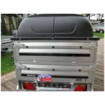 Rear view of a silver Brenderup car trailer with a black top cover, metal latches, taillights, and a dealer sticker on the back.