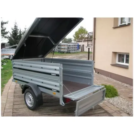 Metal utility trailer with raised lid, empty bed, and drop-down rear ramp on a brick driveway beside a beige house.