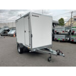 White enclosed cargo trailer on a single-axle chassis with hitch, parked in a lot with other trailers nearby.