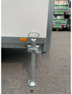 Trailer jack leg supporting a white cargo trailer, with locking pin and base plate on the ground; trucks visible in the background.