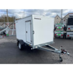 White enclosed cargo trailer with a side access door, mounted on a single-axle chassis with a tow hitch and jockey wheel, parked in a lot; brand Brenderup visible on the front corner.