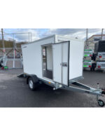 White enclosed utility trailer with a side door open and the rear ramp/door ajar, hitched to a tow bar, parked in a dealership lot.
